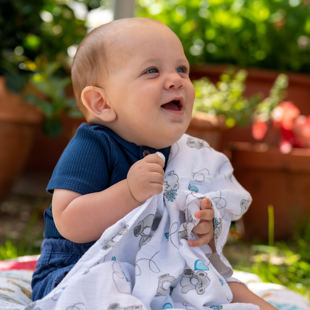 Baby sitzt lachend draußen und hält eine Tabalino Mullwindel Teddy & Wolken
